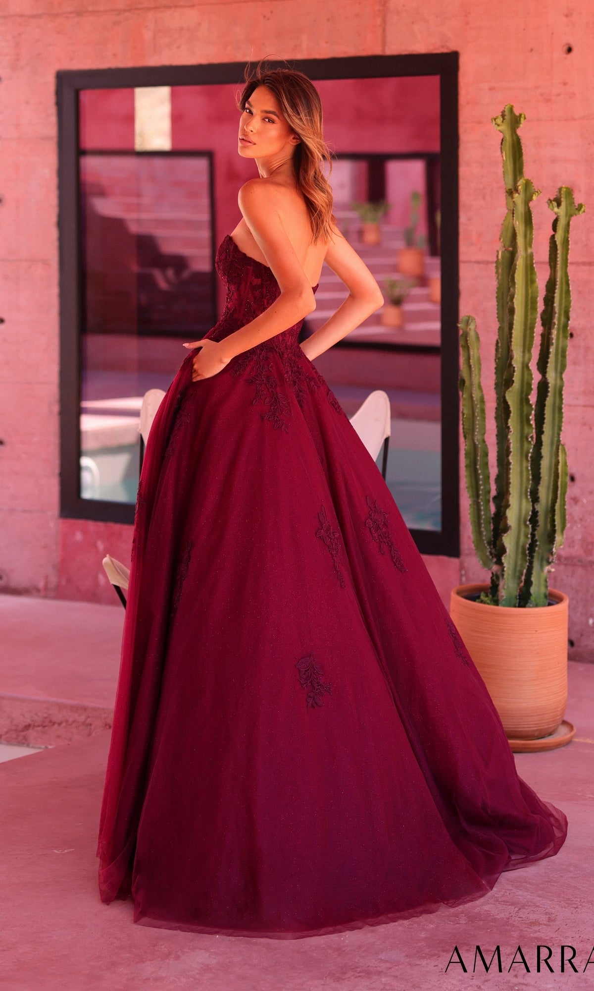 A woman wearing the Amarra 89688 Long Prom Dress—a strapless, floor-length burgundy gown—stands indoors near a tall cactus and a pink wall, with room details reflected in the window behind her.